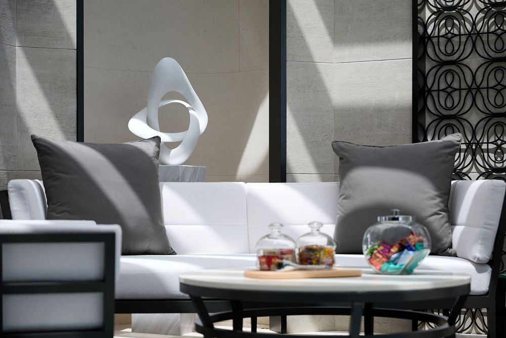 Modern outdoor lounge with white cushioned chairs, gray pillows, abstract sculpture, and glass jars on a round table.