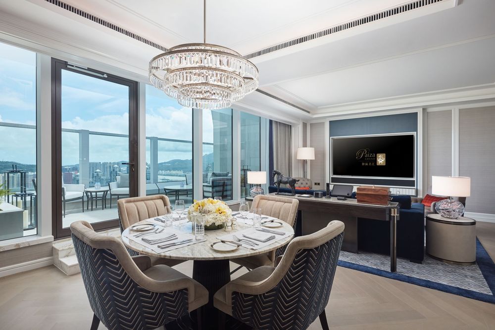 Elegant hotel dining room with round table, crystal chandelier, gray chairs, and panoramic city views through floor-to-ceiling windows.