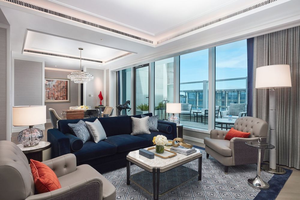 Elegant hotel suite living room with navy blue sofa, gray armchairs, and floor-to-ceiling windows overlooking a city skyline.