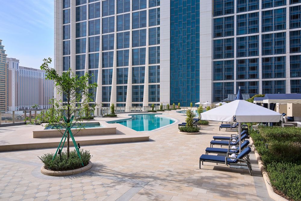 Outdoor hotel pool surrounded by lounge chairs, umbrella, and lush planters with modern high-rise buildings in the background.