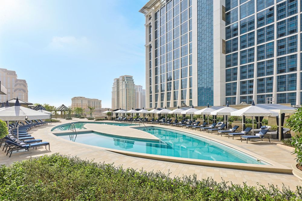 Luxury hotel rooftop pool with lounge chairs and umbrellas overlooking a modern city skyline on a sunny day.
