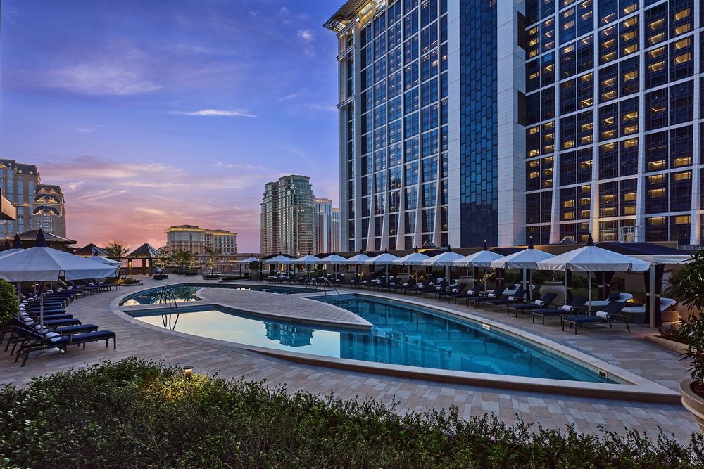 Luxury hotel pool with lounge chairs and umbrellas at dusk, reflecting city skyline in calm blue water.