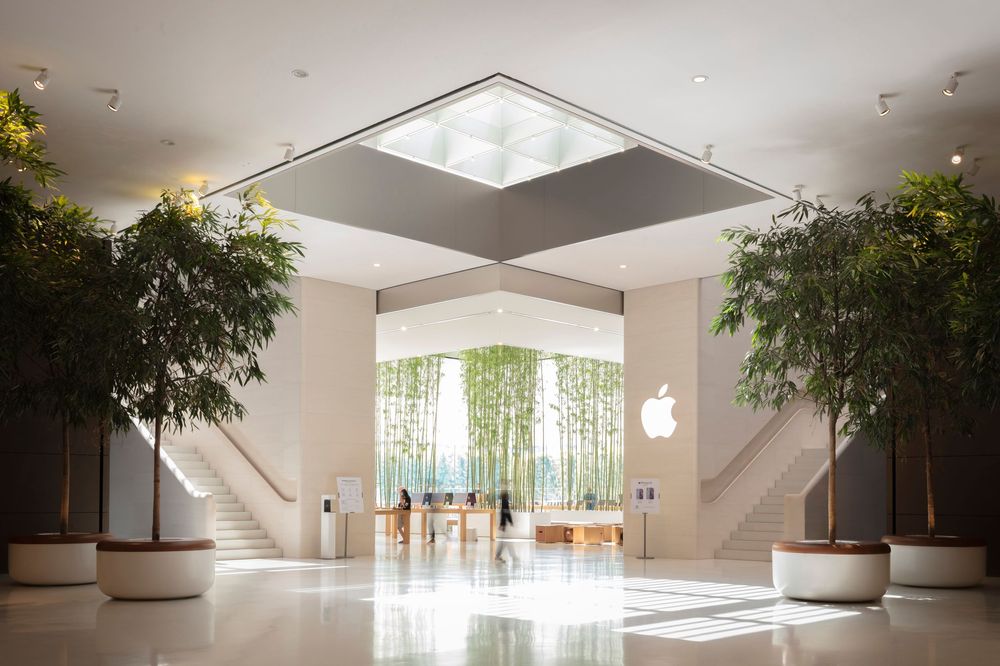 Bright Apple Store entrance with lush green trees, white floors, and iconic Apple logo glowing above product displays.