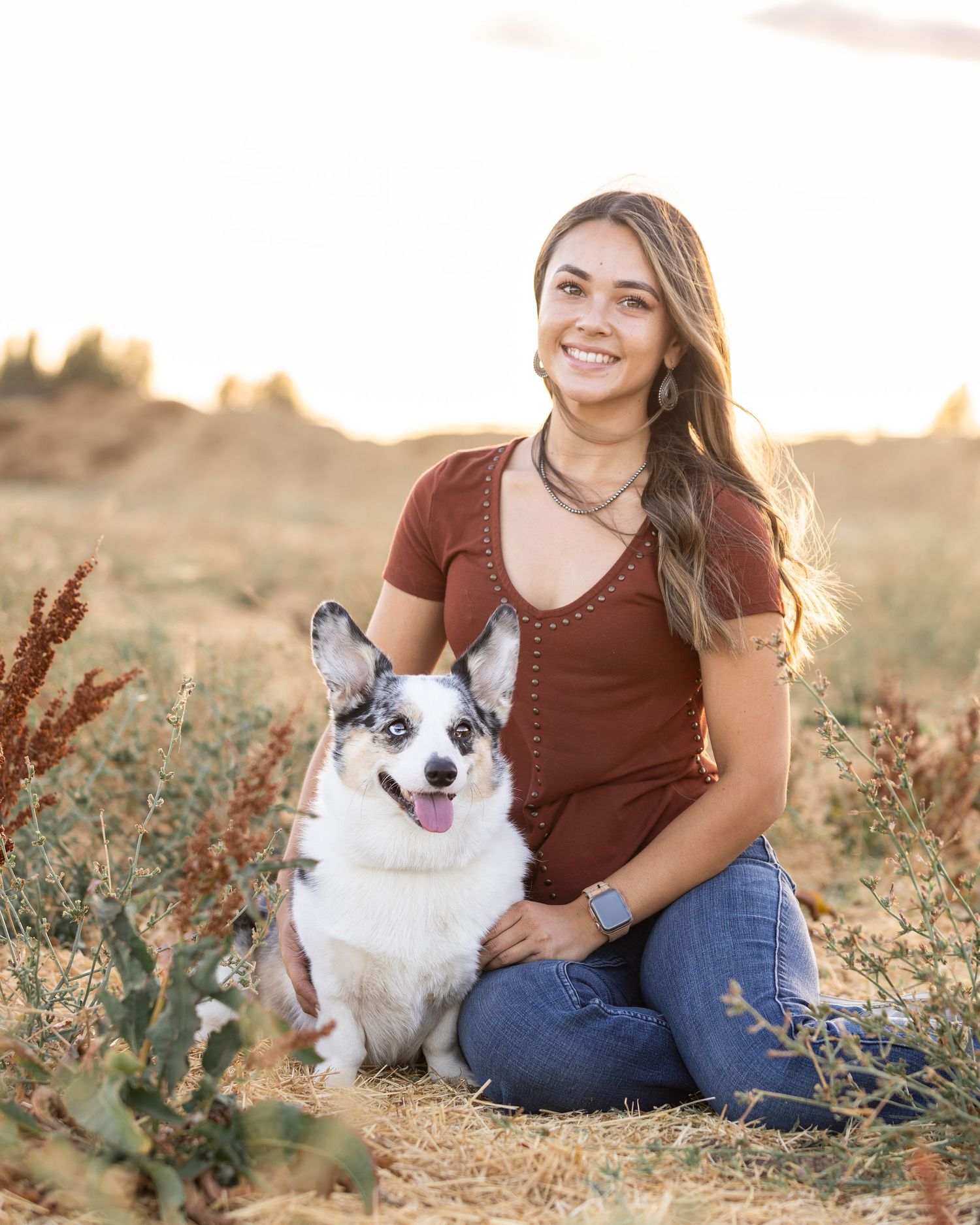 Smiling young woman in brown shirt sitting with her blue merle corgi dog in a golden field.