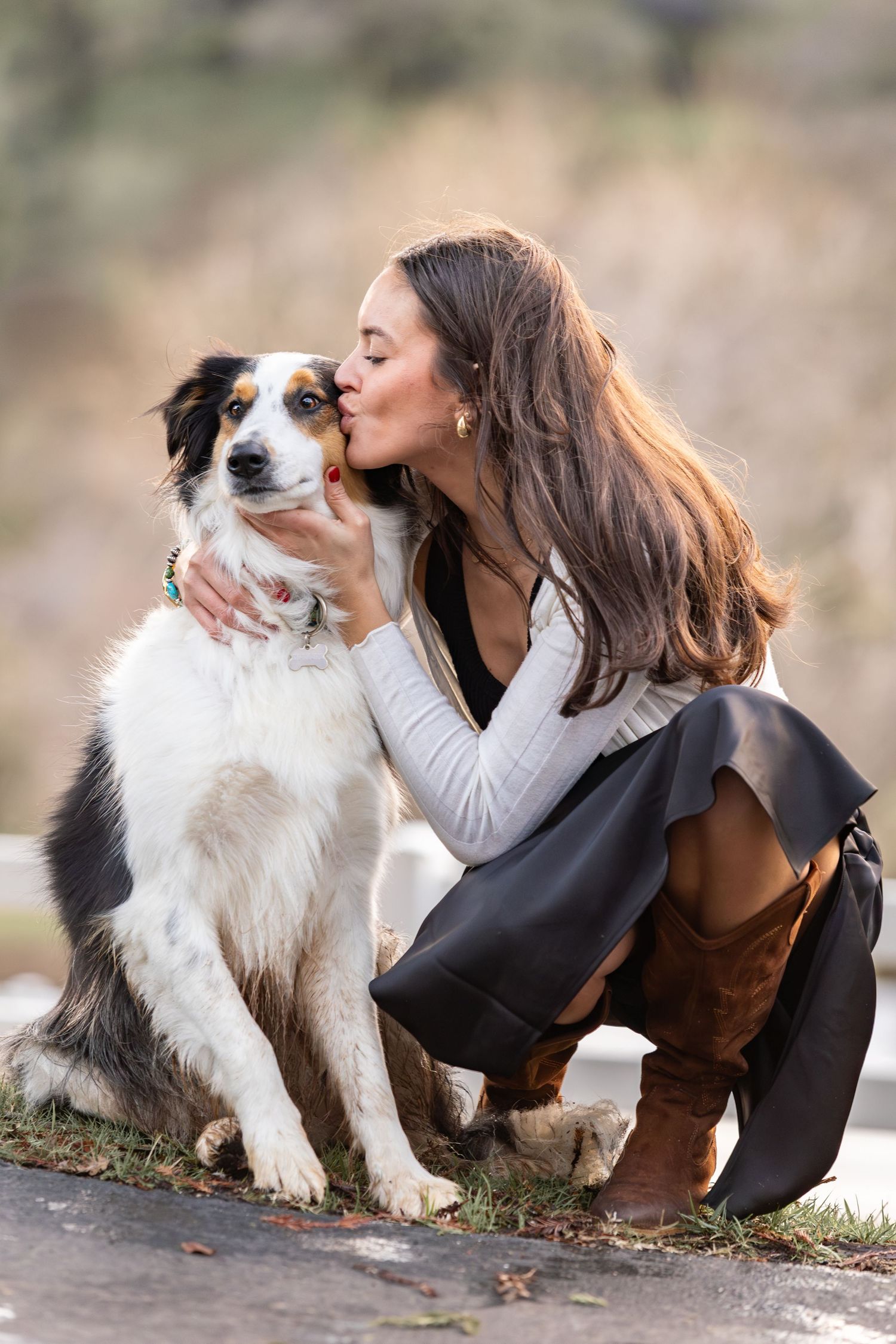Young woman in black skirt and boots kissing her Australian Shepherd dog outdoors.