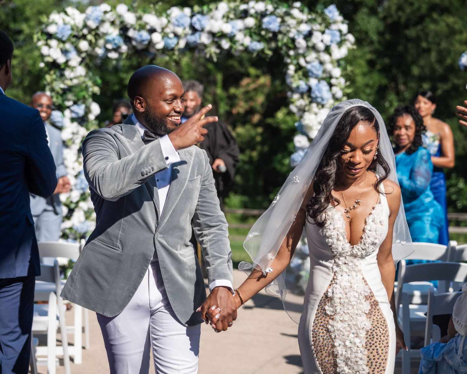 A joyful newlywed couple walks hand in hand at an outdoor wedding ceremony with blue floral arch.