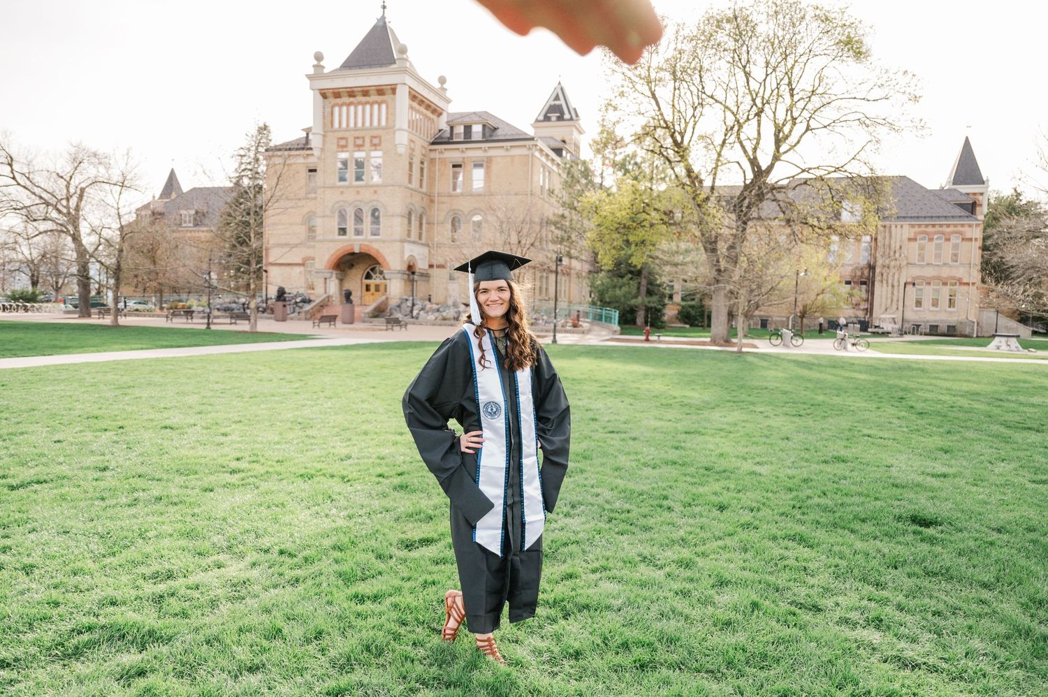 Graduate in cap and gown poses on lawn in front of grand stone university building.