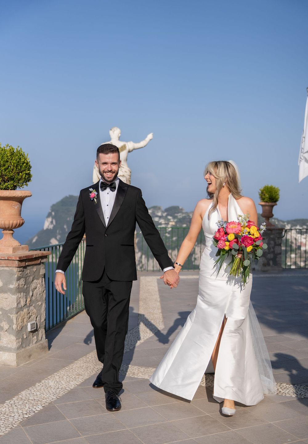 A couple in wedding attire walking and holding hands on a scenic outdoor terrace overlooking a city view.