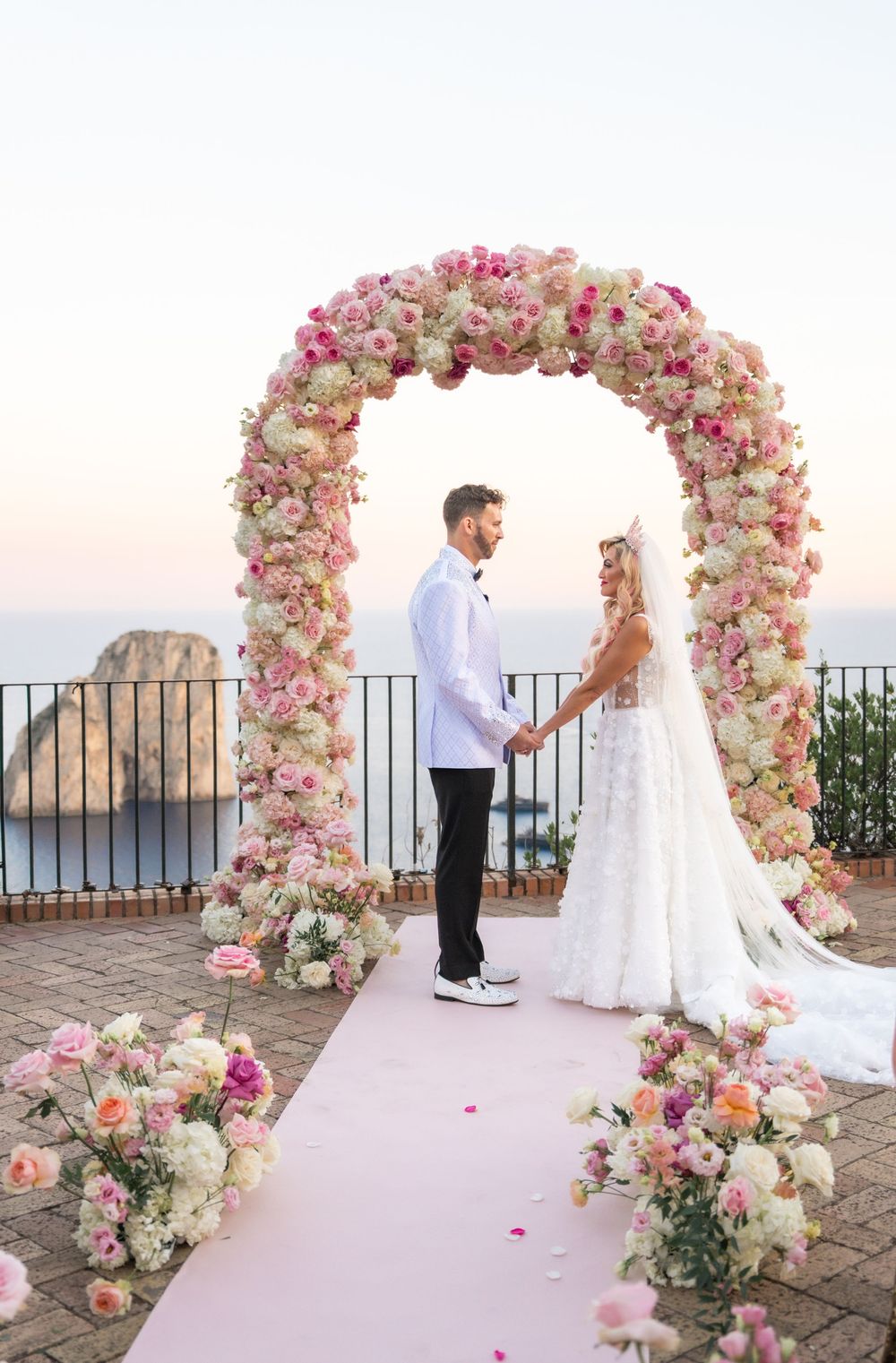 Beautiful floral arch with pink and white roses creates a romantic Mediterranean wedding ceremony backdrop at sunset.