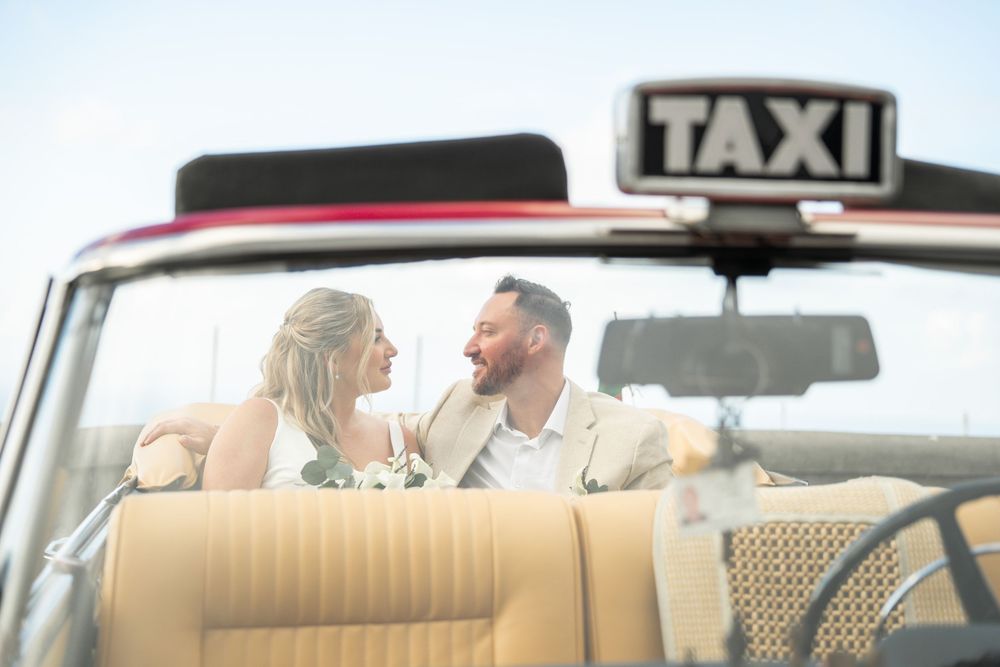 A taxi driver and passenger sit in a car with beige leather seats while driving through a city.