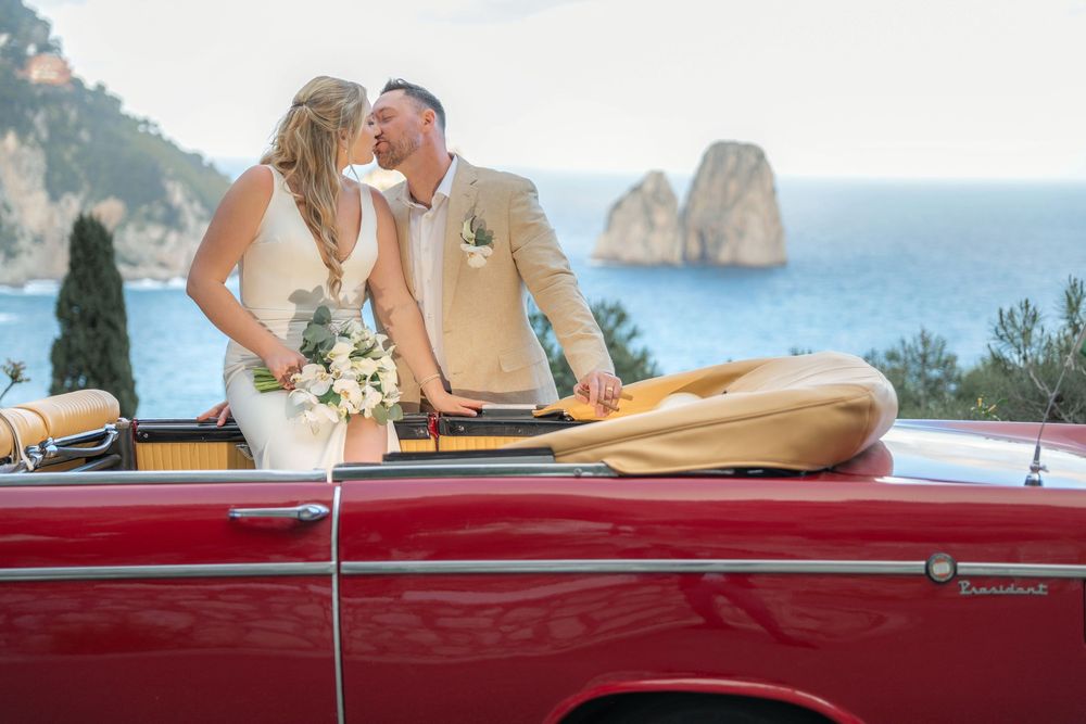 Red convertible classic car parked with coastal cliffs and blue Mediterranean waters in the background.