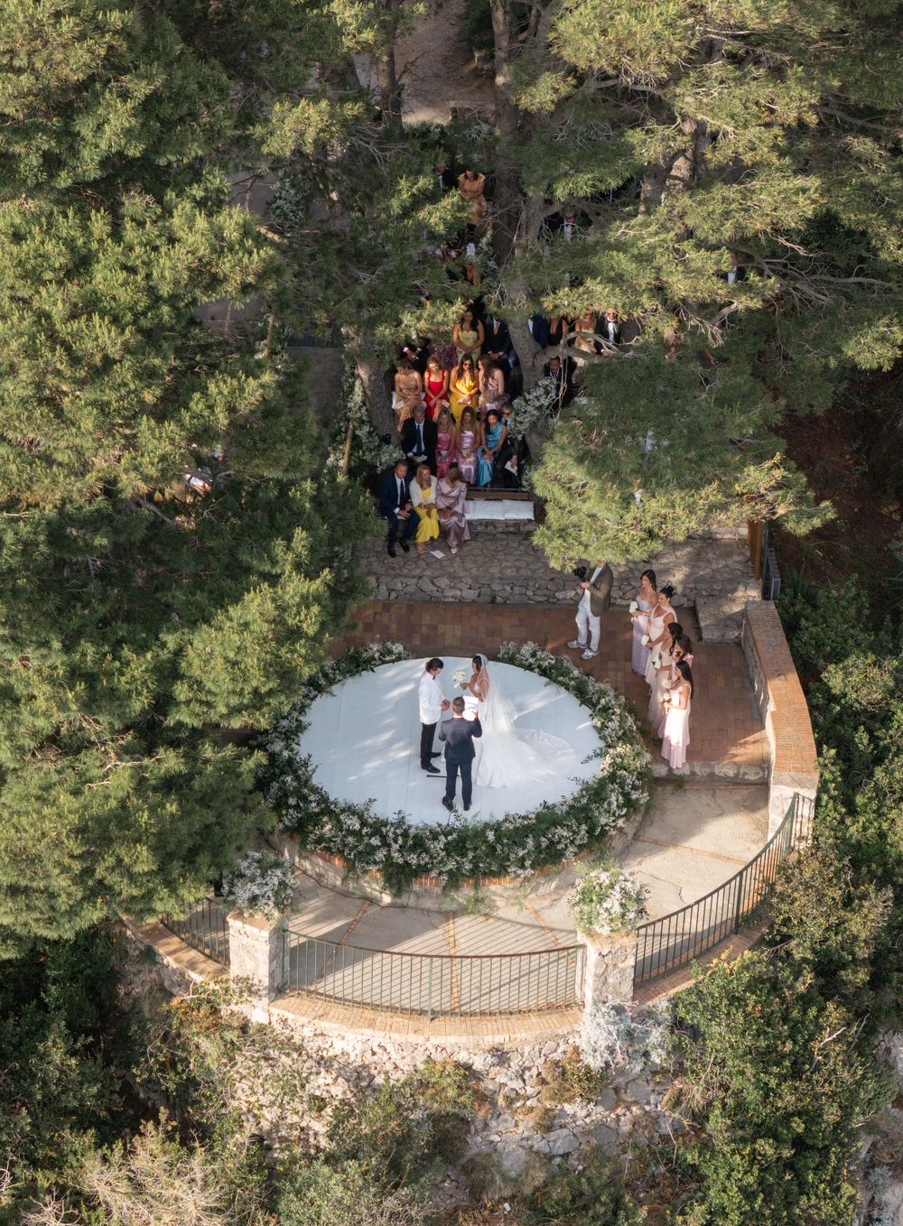 Aerial view of an outdoor wedding ceremony with a circular floral arch and guests gathered on a wooded hillside path.