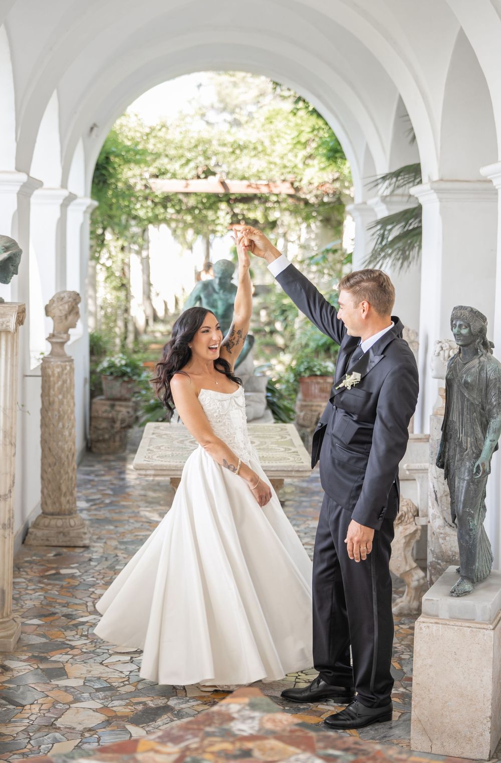 A couple in wedding attire dancing and twirling under an elegant arched colonnade with stone statues and garden views.