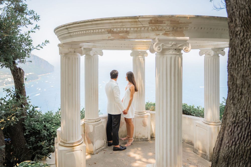A couple stands in a white columned gazebo overlooking a scenic mountain landscape on a sunny day.
