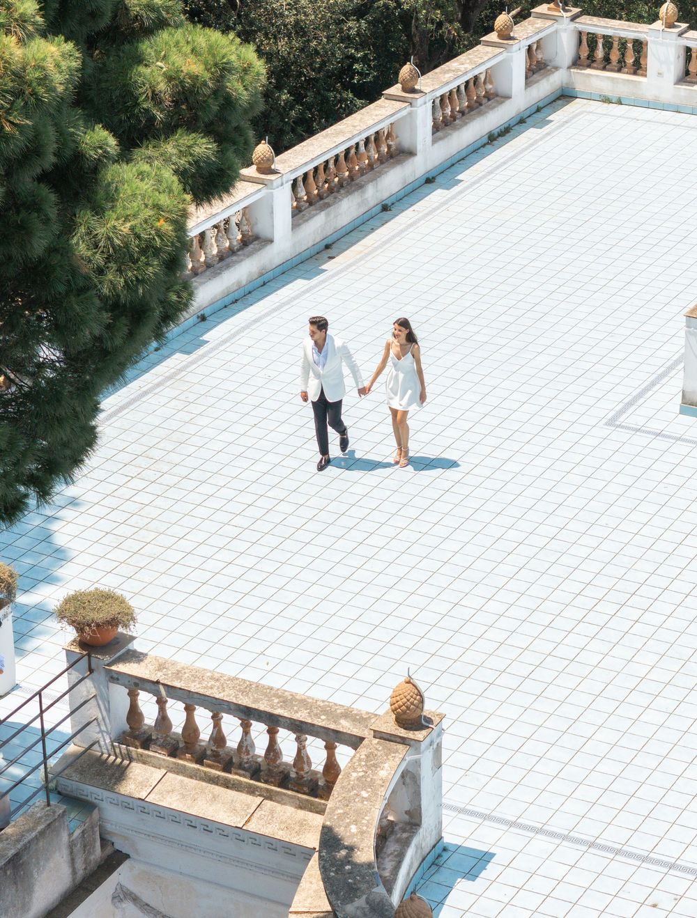 A couple shares a romantic moment on a white tiled terrace overlooking lush greenery on a sunny day.