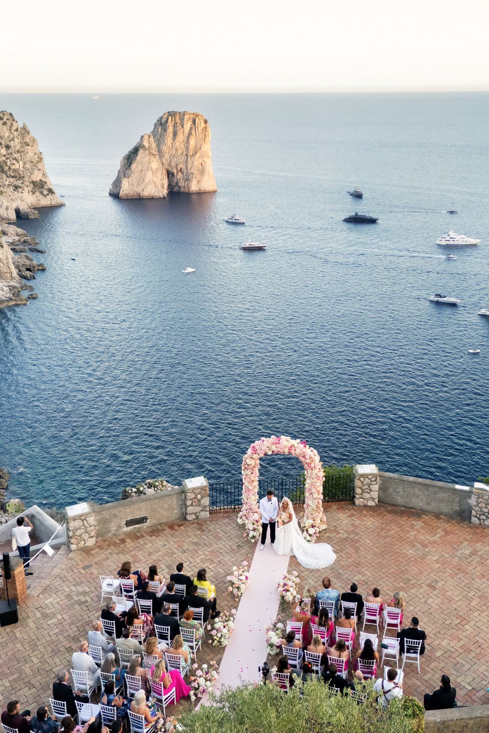 Scenic cliffside wedding ceremony overlooking the Mediterranean Sea with boats anchored near dramatic rock formations.