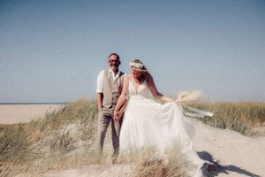 A couple stands together on a sandy beach dune with tall grass swaying in the breeze on a sunny day.
