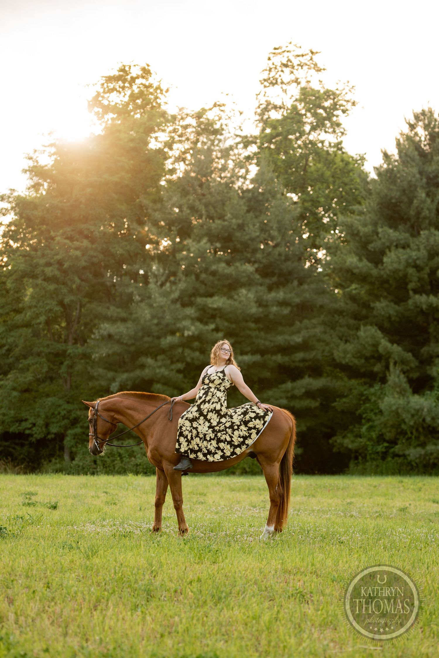 A person in a floral dress sits atop a brown horse at sunset with pine trees in the background.