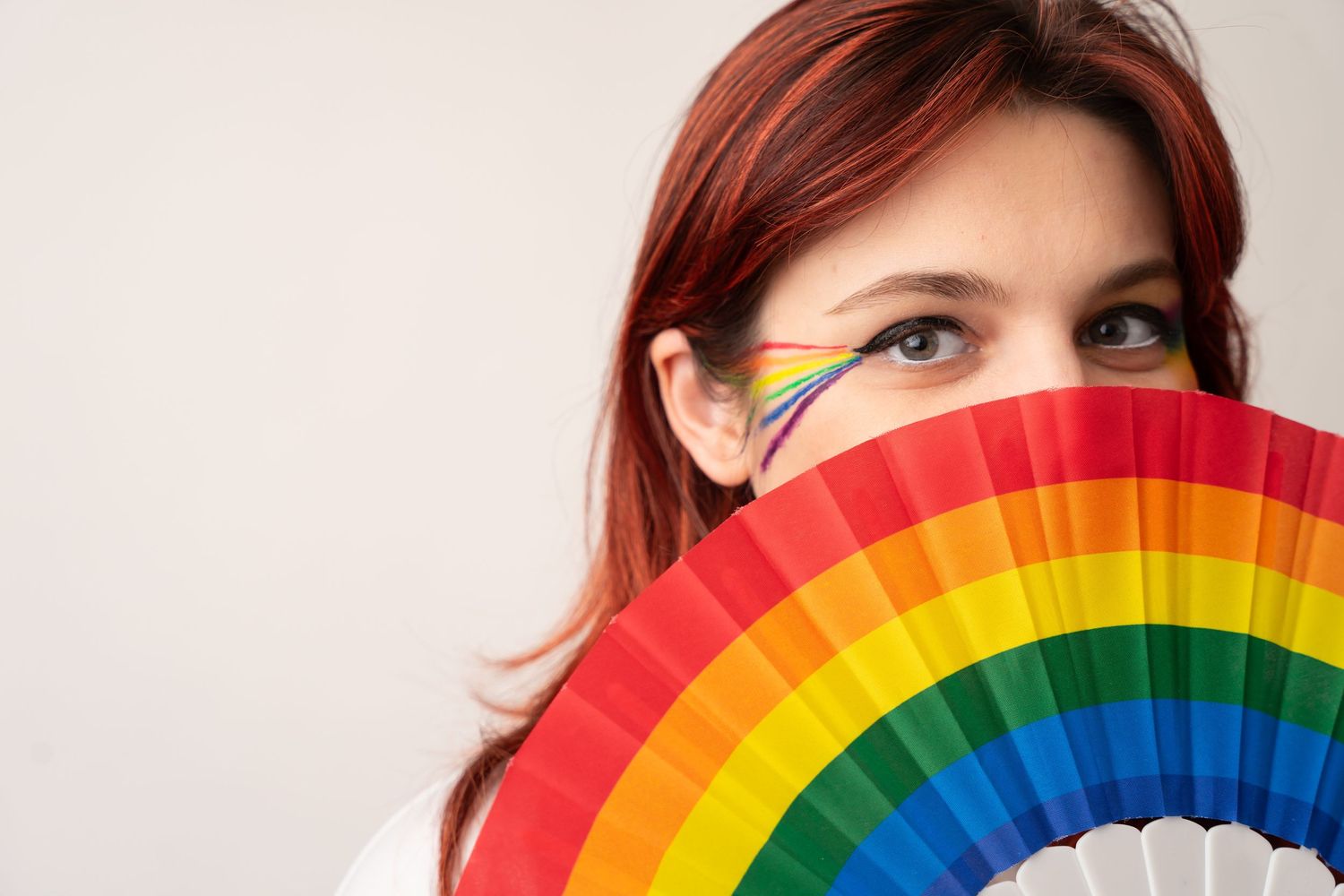 A rainbow Pride fan held up to partially cover the face against a light background.