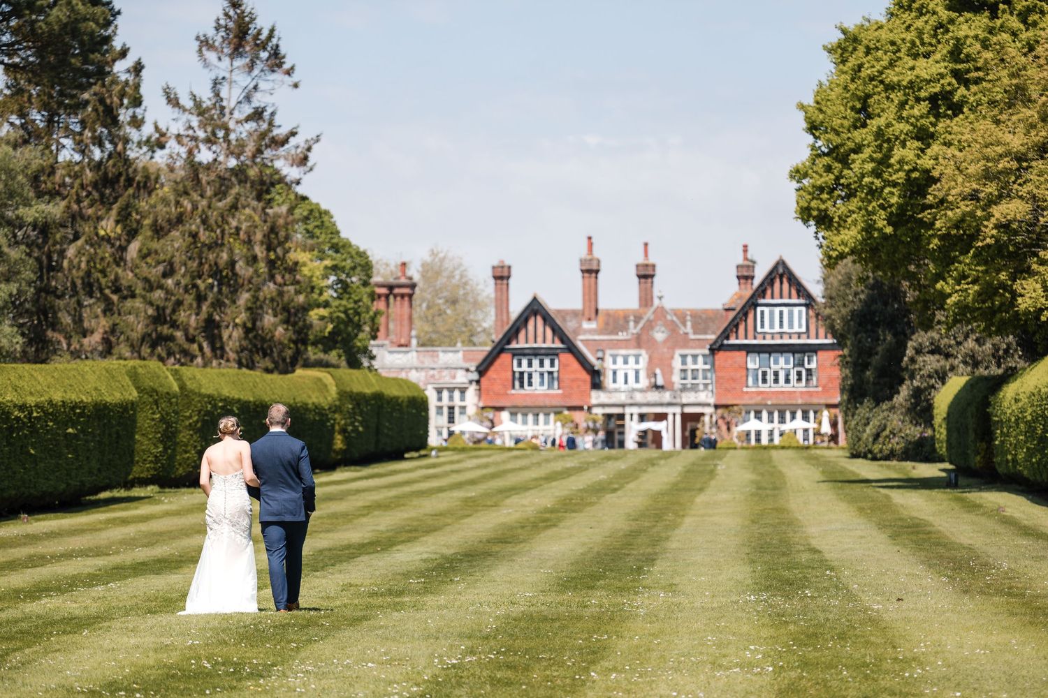 A couple walks on manicured lawn towards grand Tudor-style mansion on their wedding day.