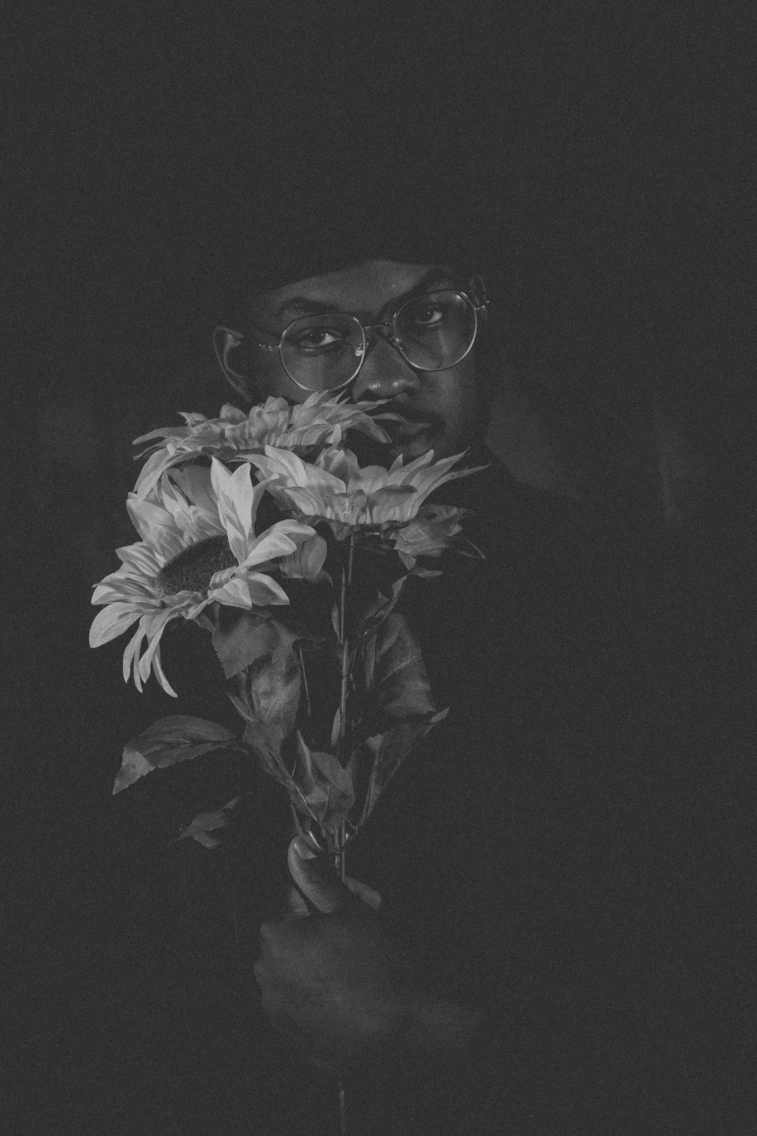 Dark moody portrait with sunflowers and glasses emerging from shadows.