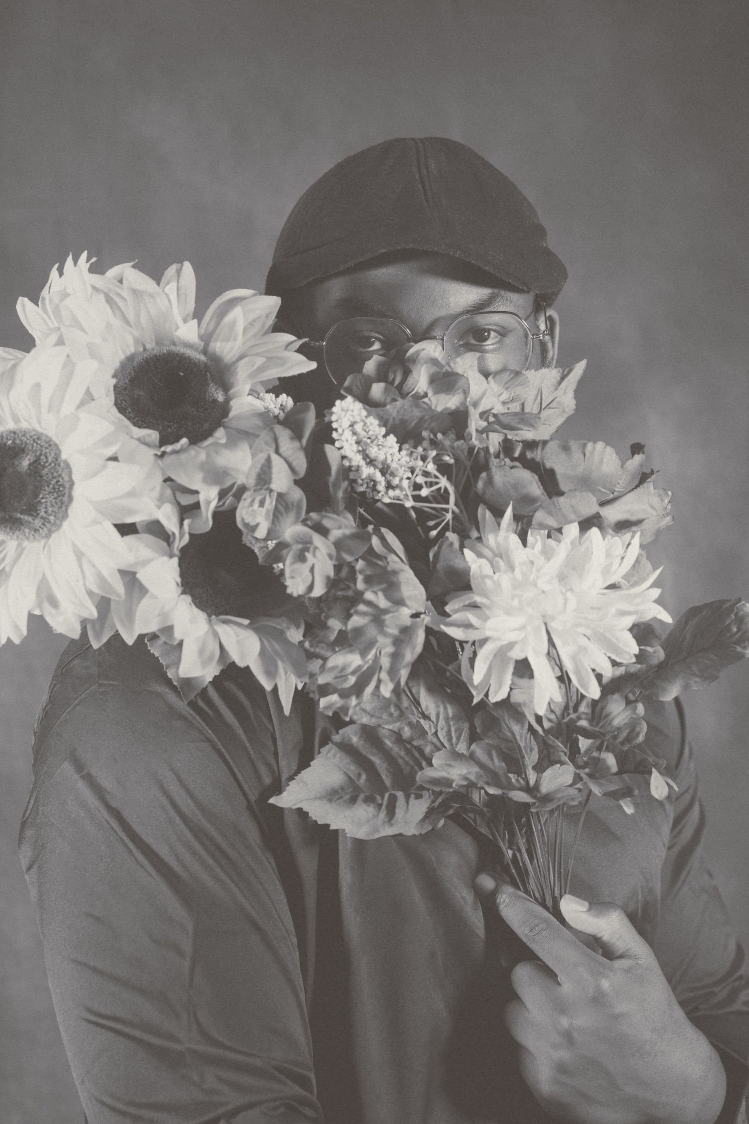 Black and white photo with sunflowers and mixed flowers held in front of subject wearing glasses.
