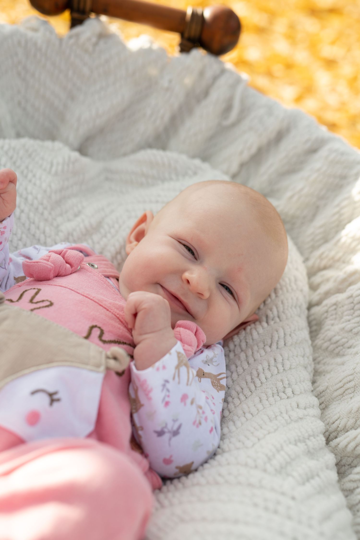 Baby in pink outfit lying on white blanket smiling with hand near mouth.