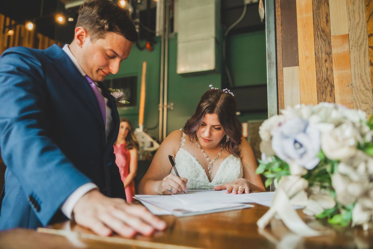 Couple signs marriage documents at wooden table with white floral arrangements.