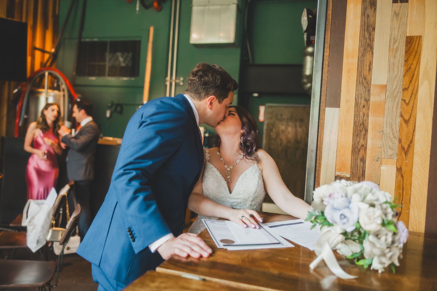 A couple shares a romantic moment while signing documents at a wooden table with a white bouquet nearby.