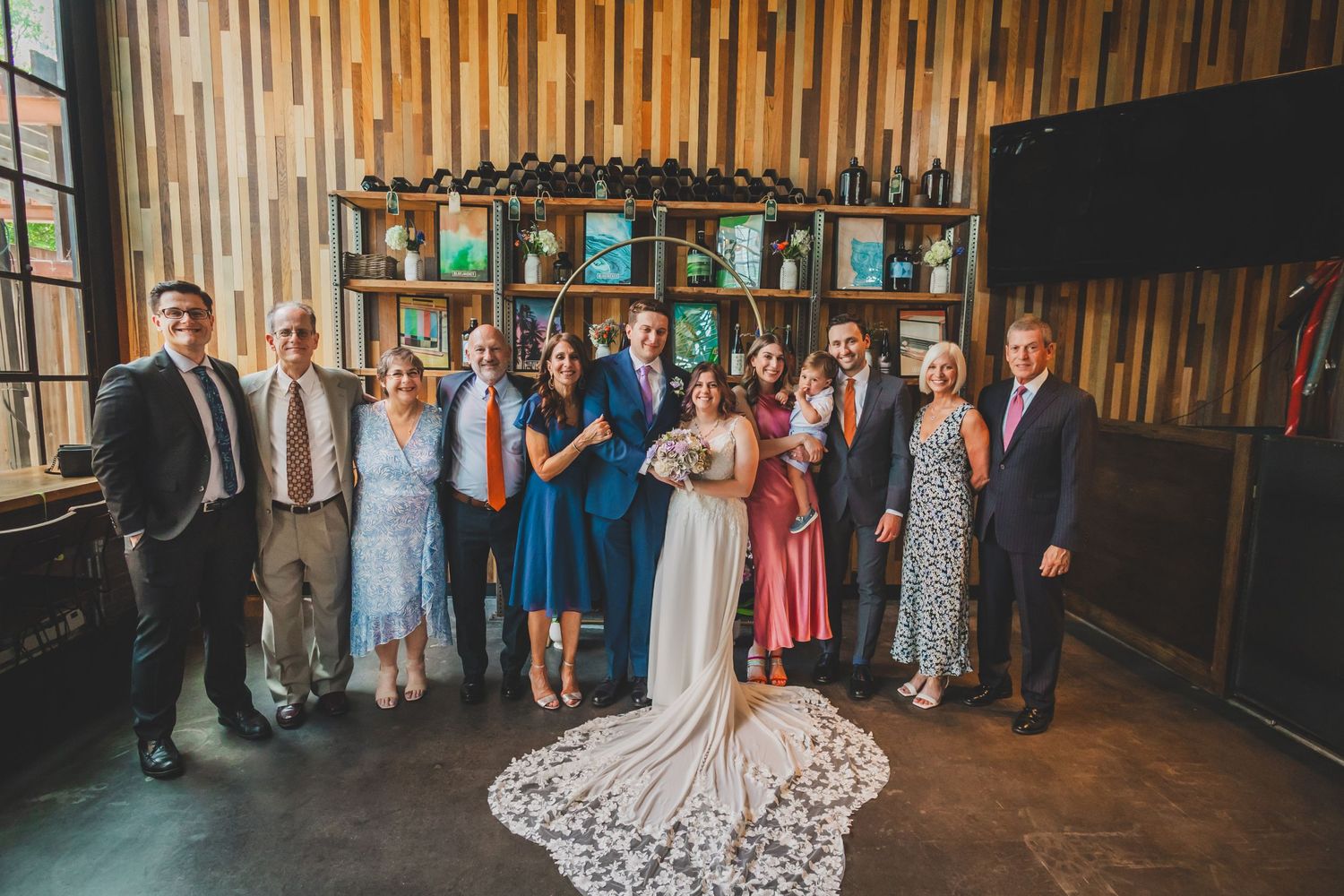 Wedding party poses in front of wooden wall decor with a long white lace train in the foreground.