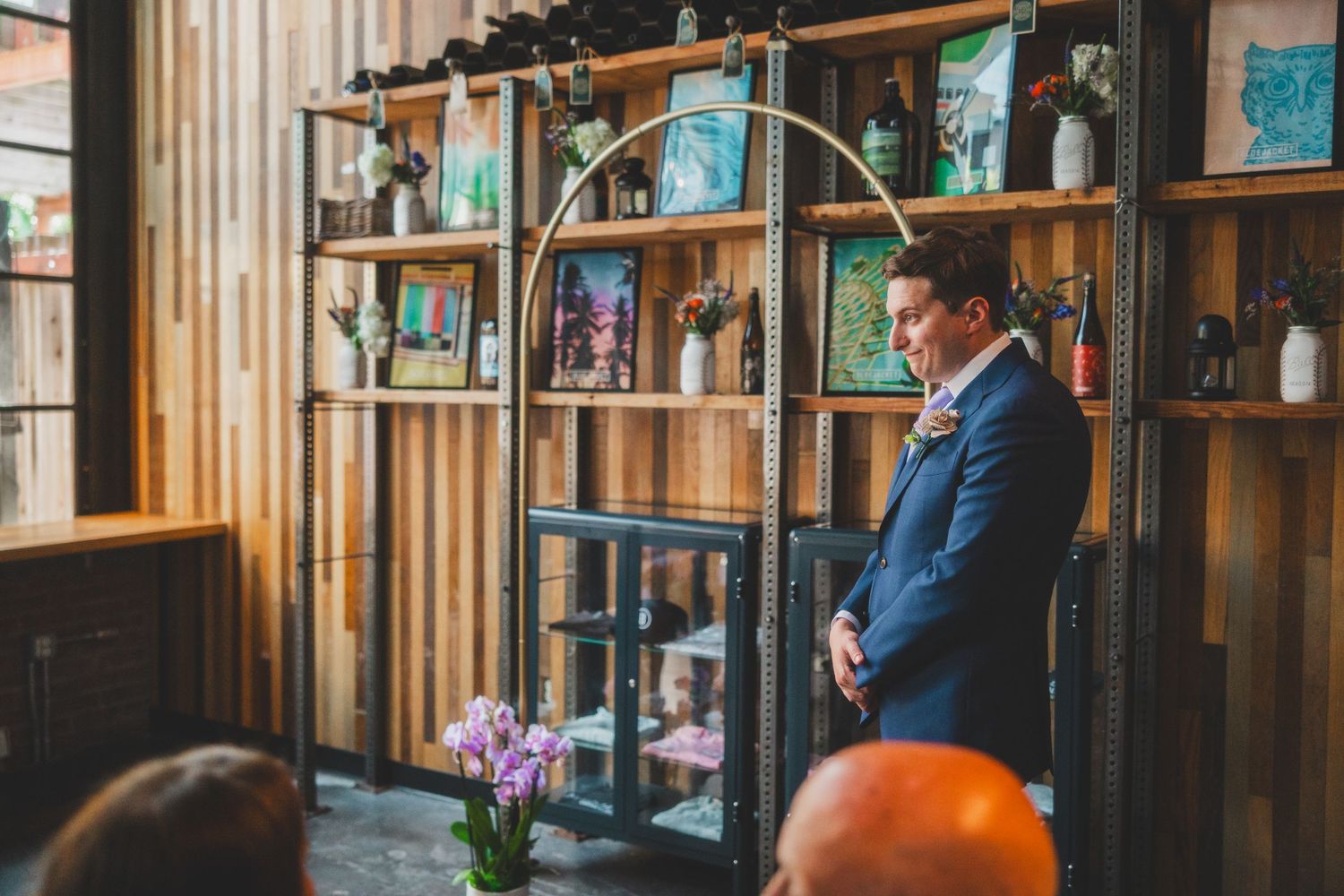 Person in blue suit stands in front of wooden shelving during wedding ceremony.