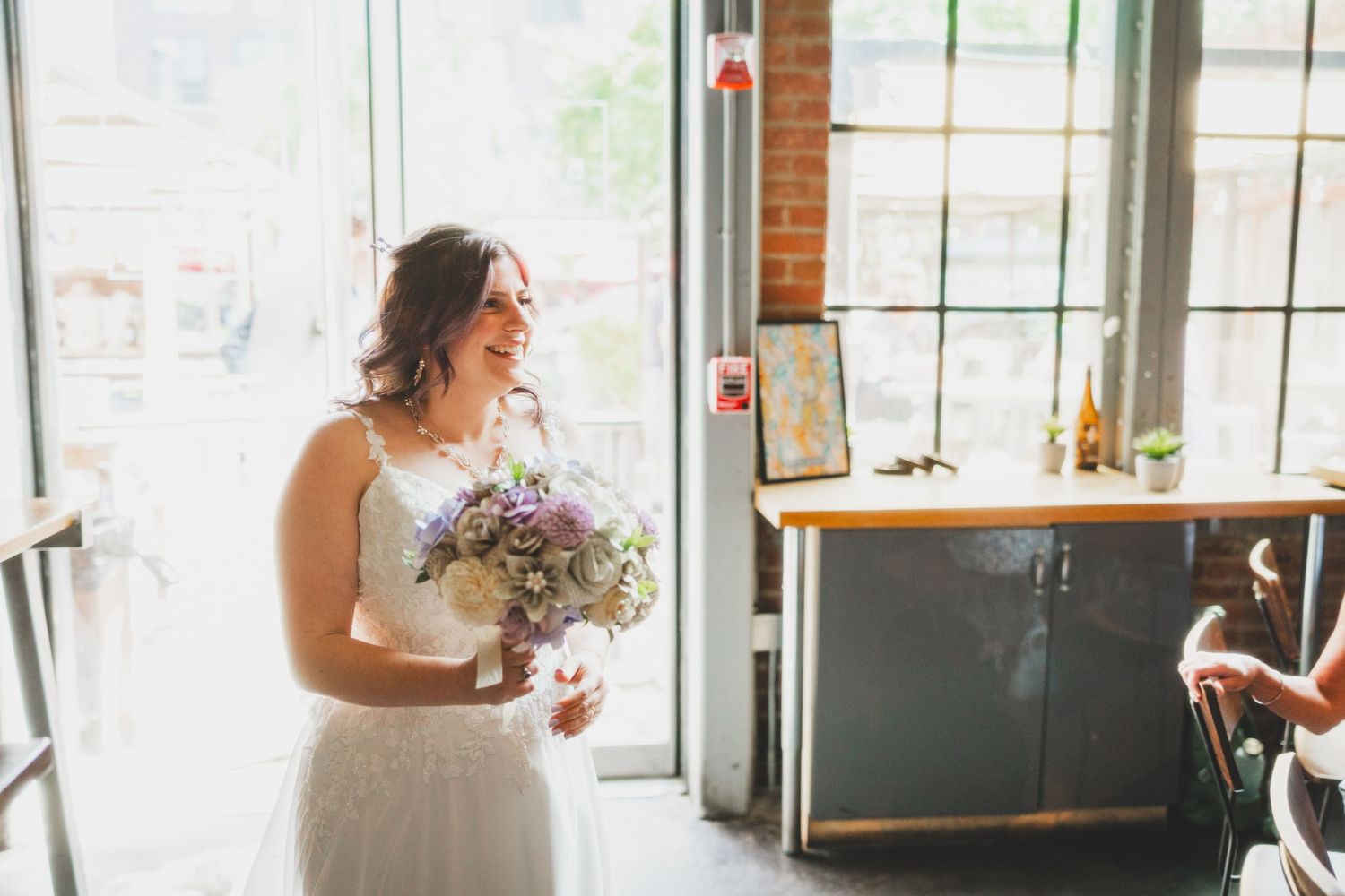 Person in white wedding dress holds purple and white bouquet near bright windows.