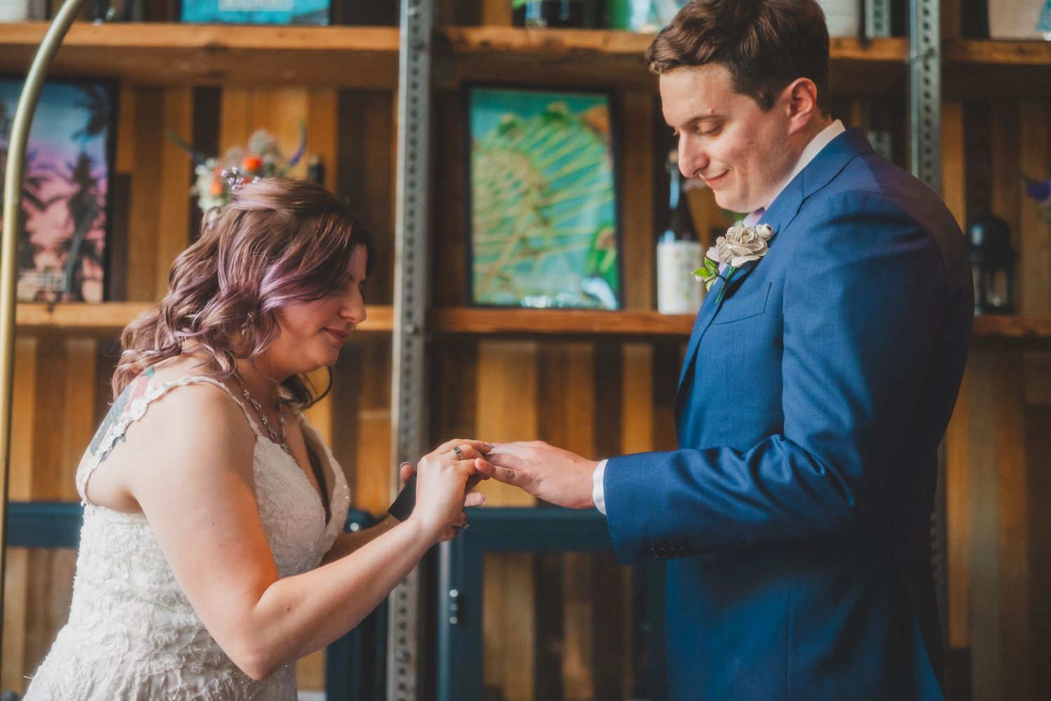 Couple exchanges rings during intimate wedding ceremony with wooden shelving backdrop.
