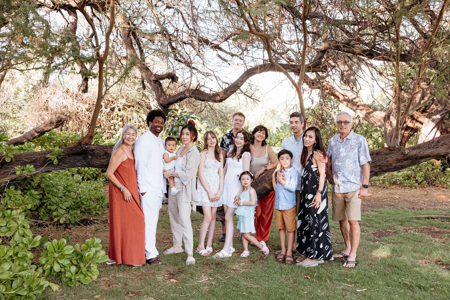 Group photo of wedding guests gathered under large banyan trees in tropical garden setting.