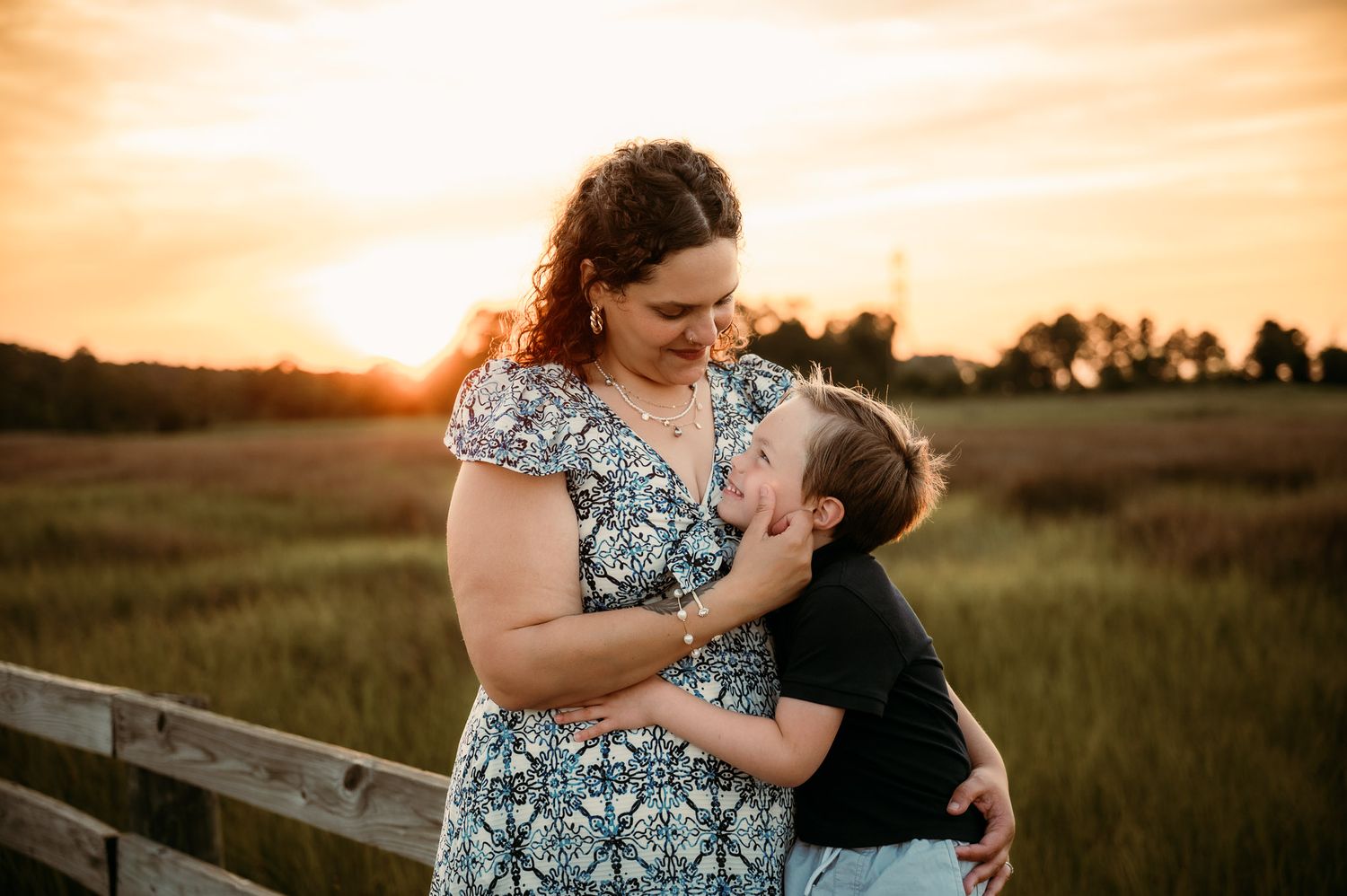 Mother and child share a tender moment at sunset in a field with wooden fence during golden hour Stafford VA.