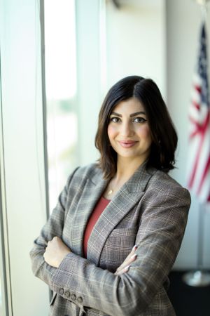 Professional in grey plaid blazer standing by window with American flag in background.