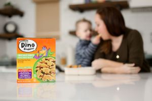 Orange box of Dino chicken nuggets on a kitchen counter with blurred figures in the background.