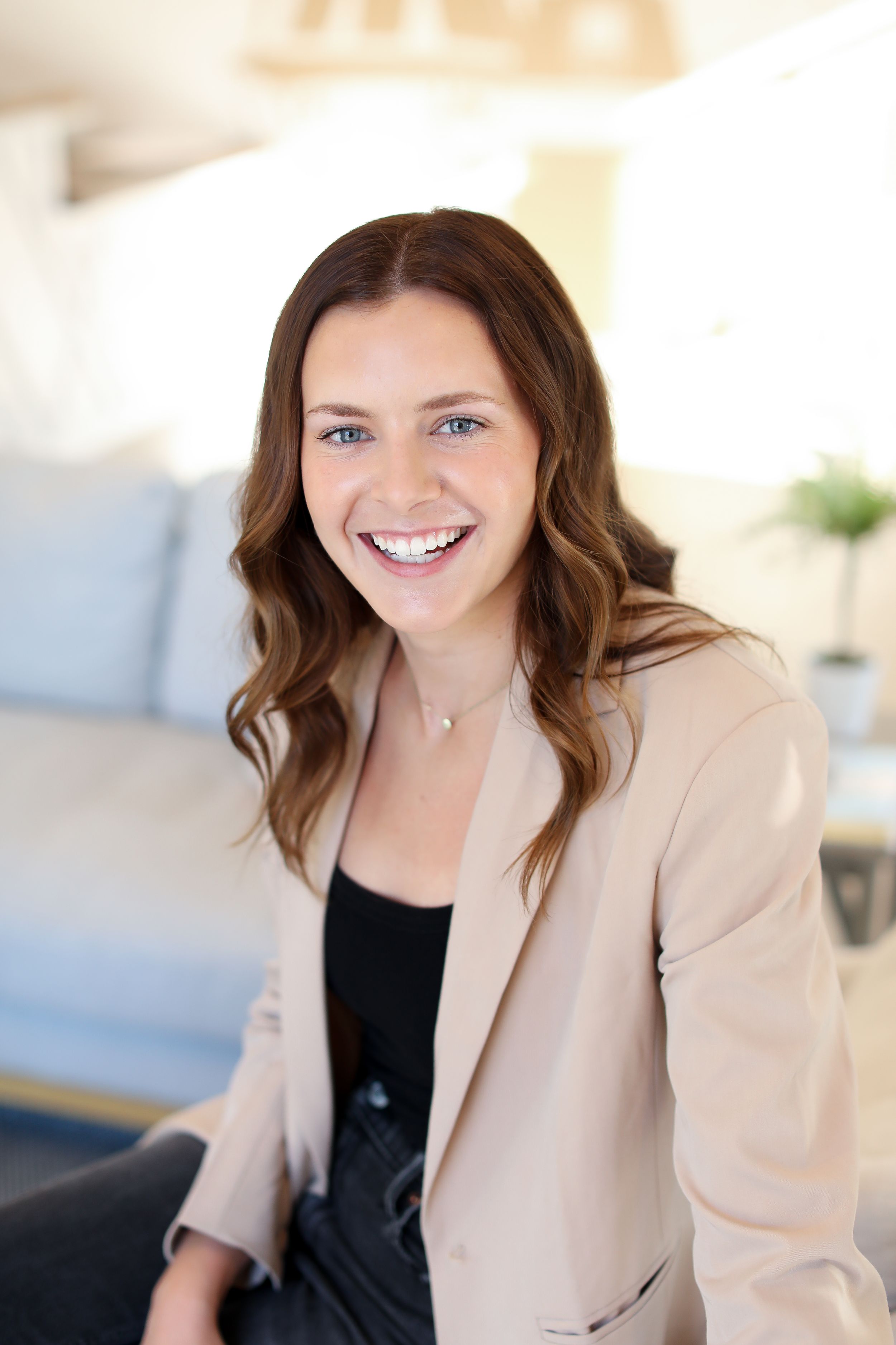 Professional headshot of someone wearing a beige blazer and black top in a bright office setting.