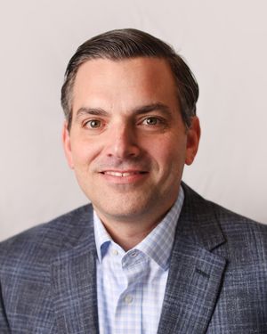 Professional headshot of a smiling executive in a grey suit and light blue dress shirt.