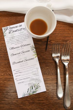 Event menu for Women in Dentistry conference placed beside silverware and coffee cup on wooden table.