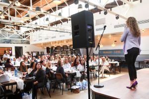 Speaker presenting to a packed room of attendees seated at round tables in a conference space.
