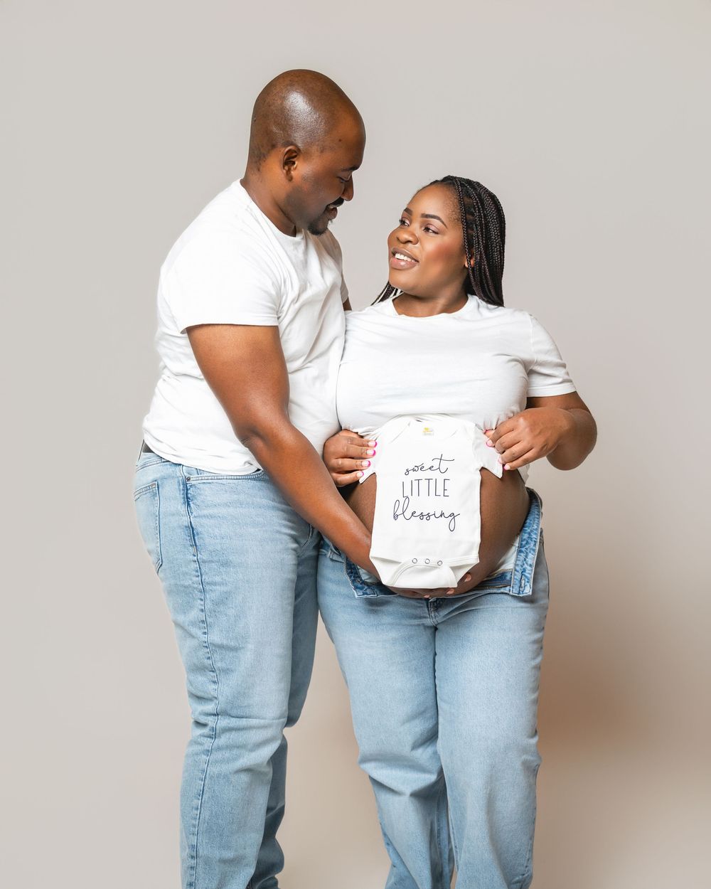 A couple in matching white t-shirts and light jeans embrace while showing their baby announcement shirts.