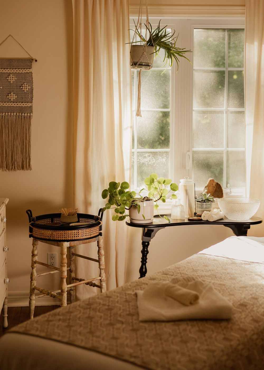 A serene massage therapy room with natural light, hanging plants, and macrame wall decor creating a peaceful ambiance.