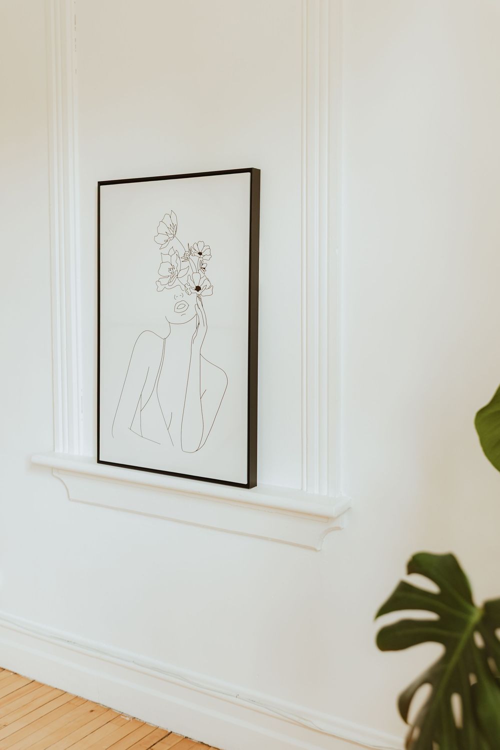 Minimalist line drawing artwork in black frame displayed on white wall with monstera plant leaf visible in corner.