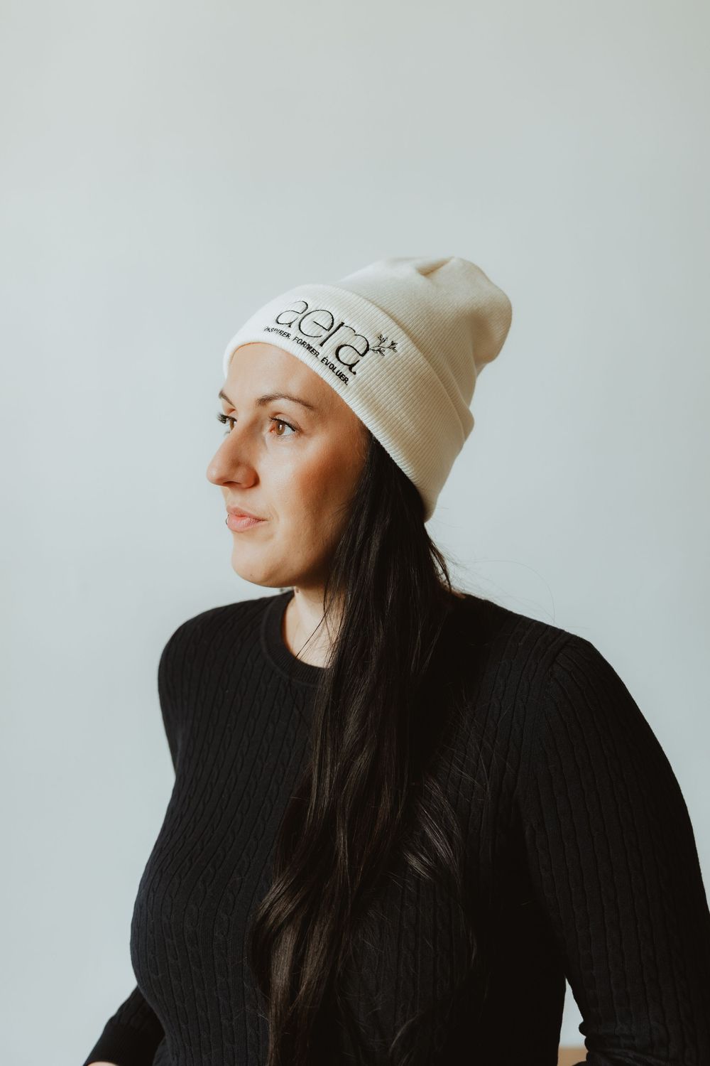 A person wearing a cream beanie and black sweater poses against a light gray background in a minimalist portrait.