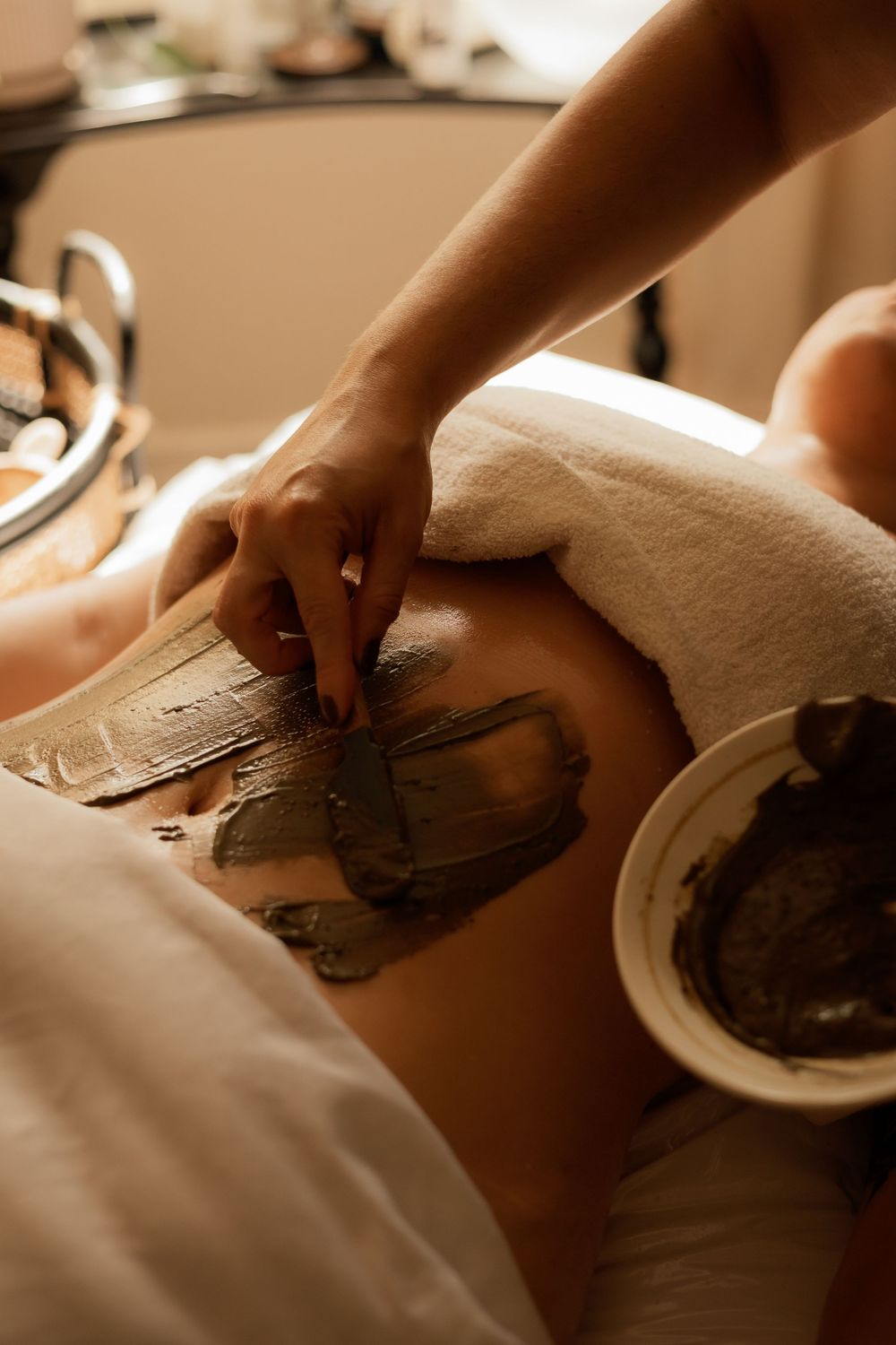Spa treatment showing application of mud or clay mask with white towels in warm ambient lighting.