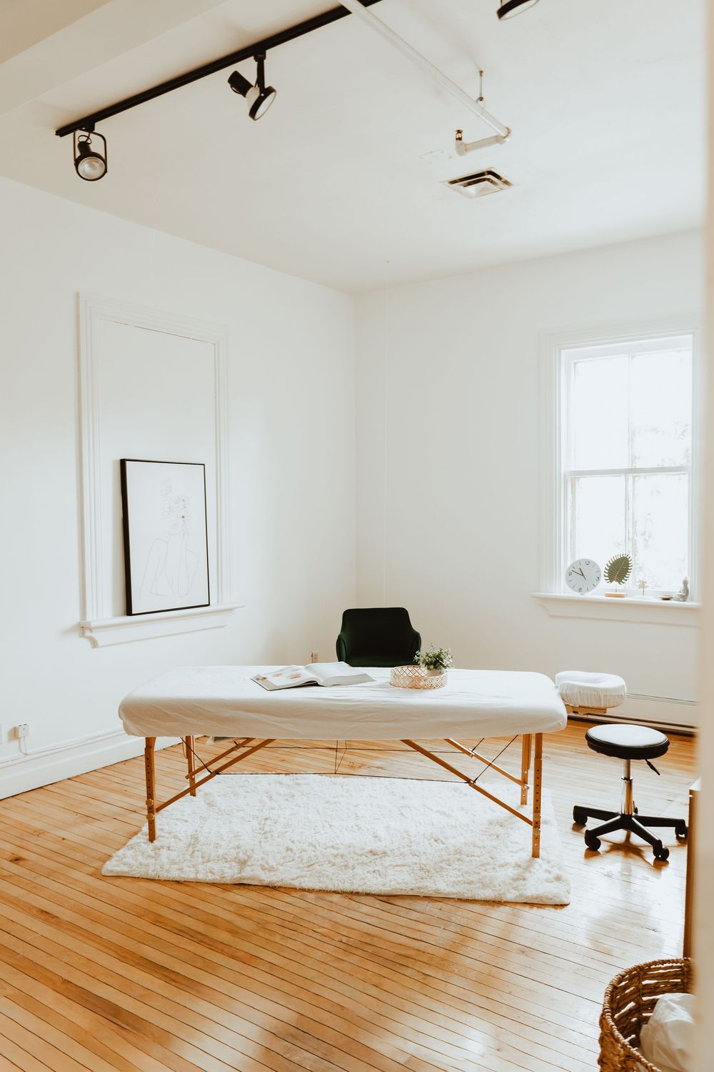 Minimalist white bedroom with wooden floors, massage table, and office chair in bright natural light.