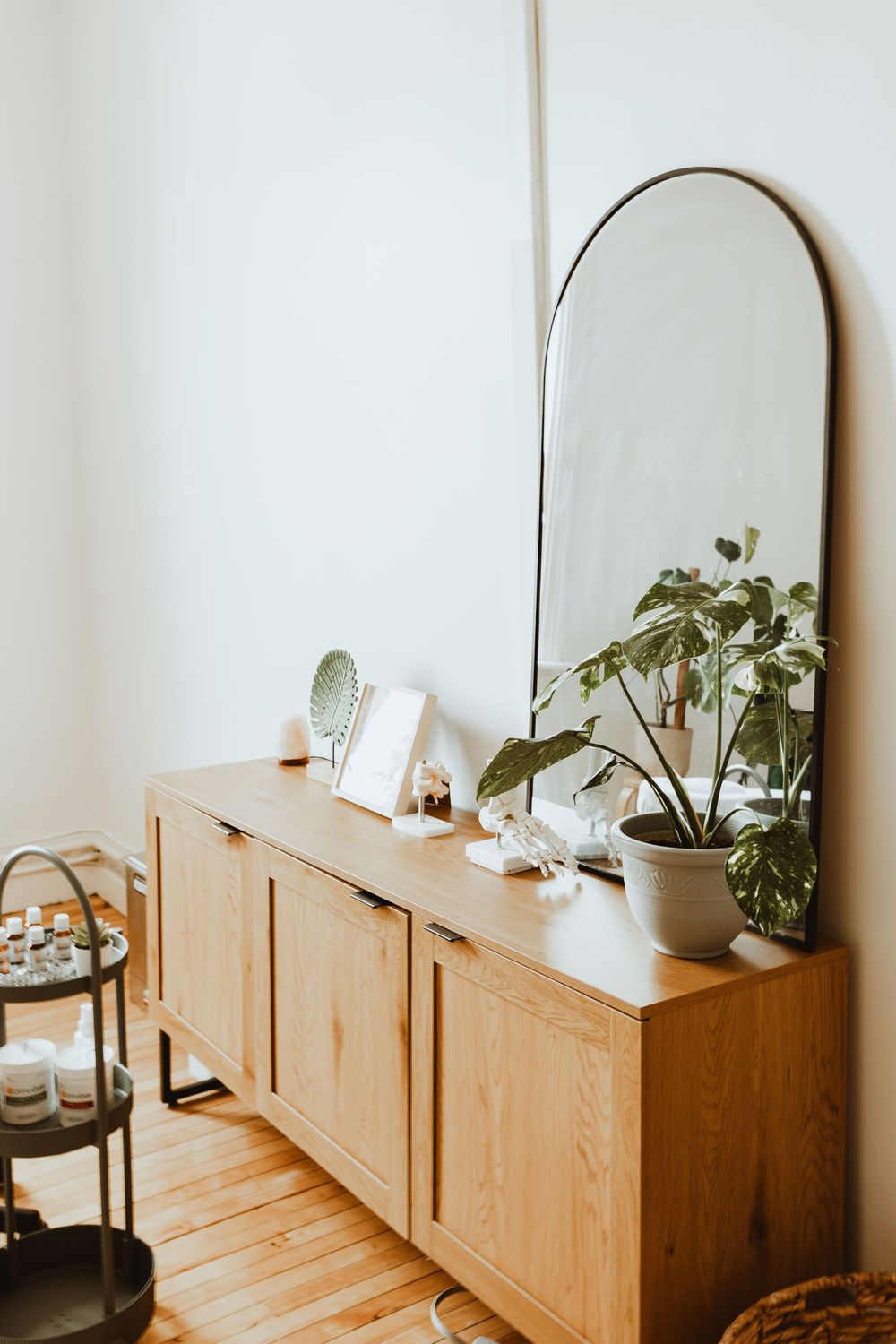 Modern wooden credenza with arched mirror and potted plants in a bright white room.