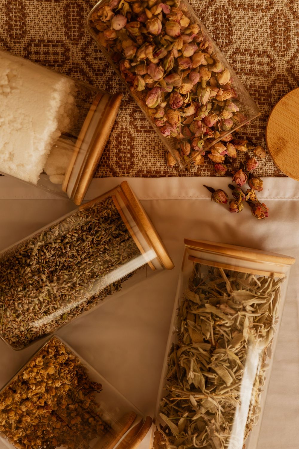 Glass jars filled with dried herbs, spices, and floral buds arranged on a textured fabric surface in warm lighting.