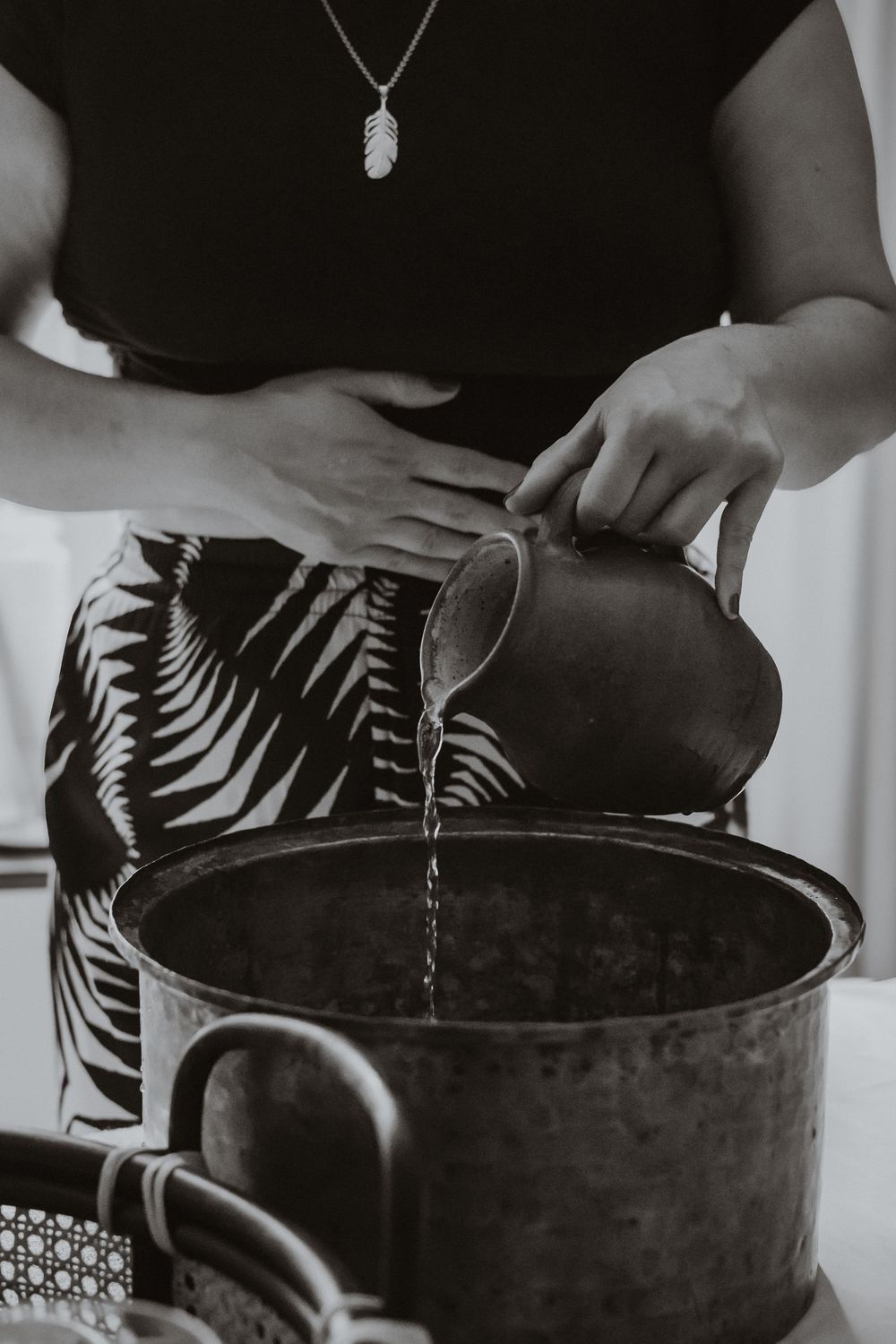 Hand pouring water from black ceramic kettle into pot against dark background with silver necklace.